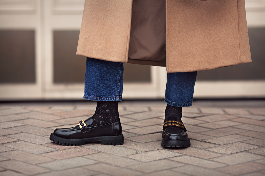 A close-up of a man wearing black loafers, jeans and a long brown coat.